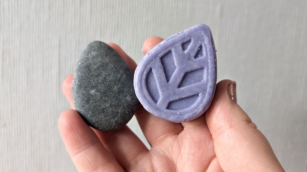 Two ecoliving solid shampoo bars being held up. The one on the left is grey and has been used for 4 weeks, the one on the right is purple and is new.
