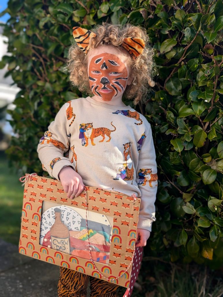 A child in a Tiger Who Came to Tea themed outfit with handmade DIY props of food and crockery items all stored in a handmade reused carboard envelope. The bag has been decorated and covered with colourful paper tapes from Babipur. 
