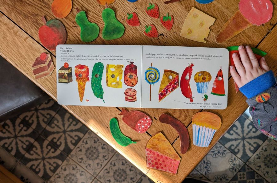 An open book - The Hungry Caterpillar, placed on a table surrounded by DIY crafted props of food featured in the book. 